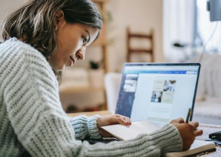 Side view of concentrated young Latin American female freelancer in casual clothes using laptop while taking notes in notebook with pen at table near ring light with phone in room