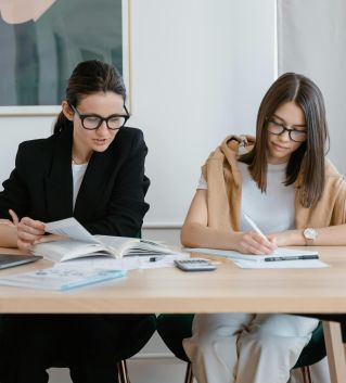 Two women working together with books and a laptop, focused on learning.